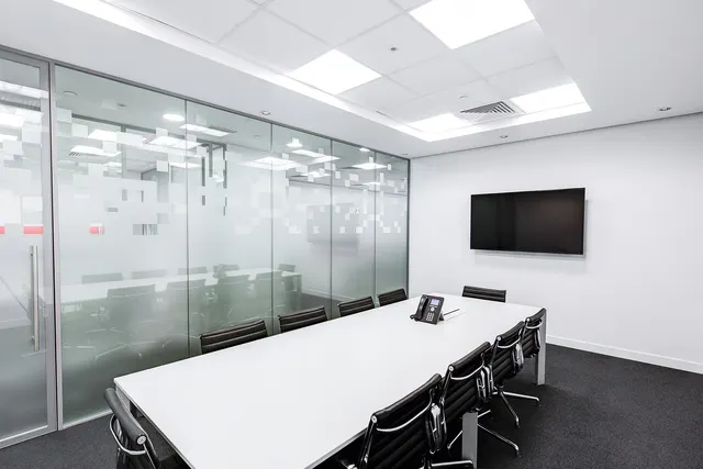 Empty modern conference room with long white table, black leather chairs, desk phone, wall-mounted TV and frosted glass wall