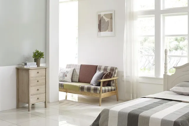 Bright, sunlit bedroom with a plaid wooden-frame sofa, light wood dresser with plant, and bed with a striped gray cover