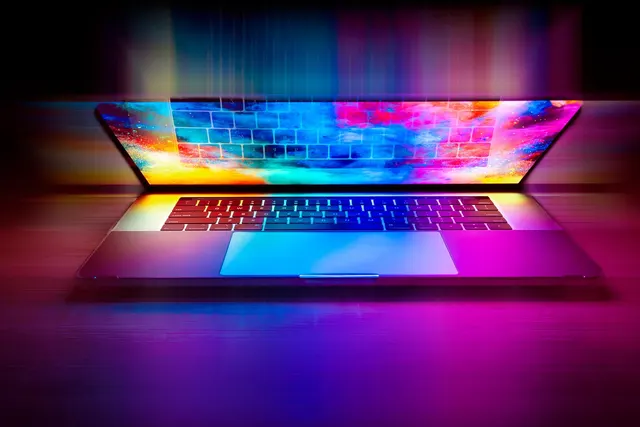 Partly open laptop on a desk with colorful light reflecting off its keyboard and screen in a dark room