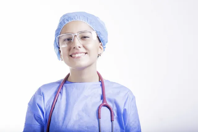Smiling healthcare worker in blue scrubs, surgical cap and safety glasses with a stethoscope around her neck