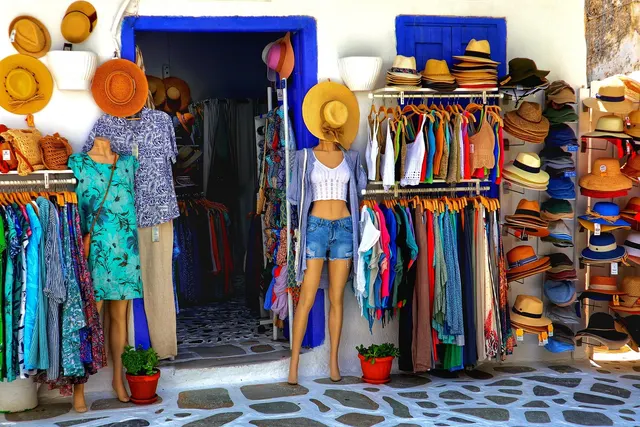 Clothing boutique entrance with racks of colorful garments, mannequins in summer outfits, and a wall of straw hats