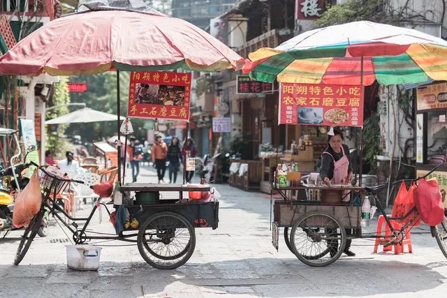Two street food tricycle carts under colorful umbrellas, one vendor in a pink apron preparing food on a sunny shopping street