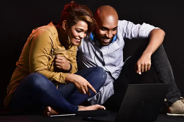 Smiling woman and man sitting on the floor pointing at a laptop screen together against a dark background