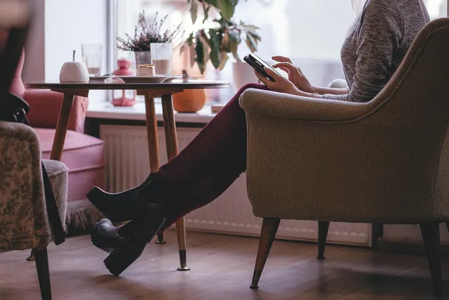 Woman in burgundy jeans and black ankle boots sits in a tan armchair by a cafe window, using a smartphone