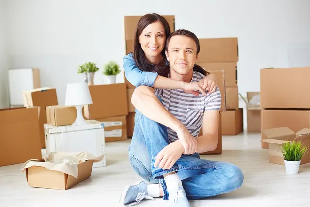Smiling couple sitting on the floor of a bright empty room surrounded by stacked cardboard moving boxes