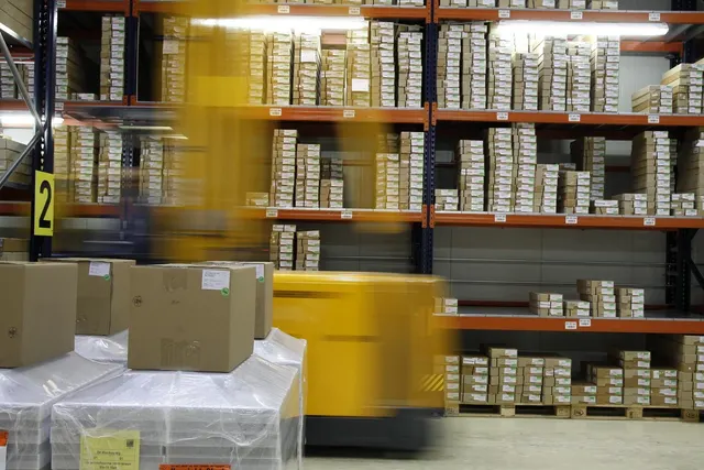 Motion-blurred yellow forklift passing tall warehouse shelves of stacked boxes beside shrink-wrapped pallets