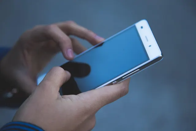 Close-up of two hands holding a white smartphone with a blank screen, thumb poised to tap, against a muted blue-gray background