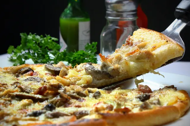 Spatula lifting a slice of mushroom and cheese pizza, with parsley, a green sauce bottle and a glass shaker behind