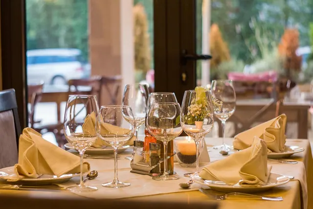 Restaurant table set with wine glasses, folded napkins on plates, a lit candle and small flowers by a window