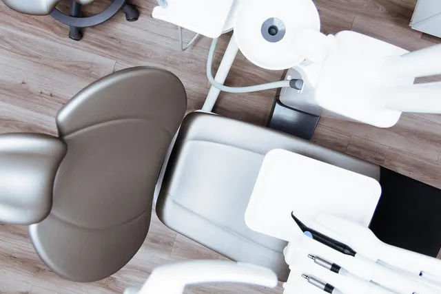 Overhead view of an empty taupe dental chair with white instrument tray and handpieces on a wood-look floor