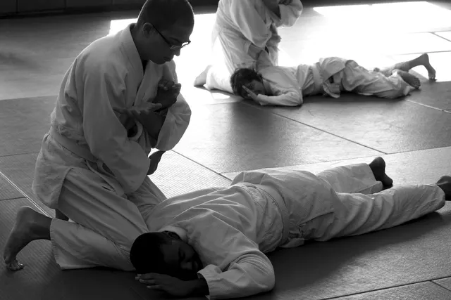 Martial arts students in white uniforms practicing an arm pin on training mats in a dojo, black and white