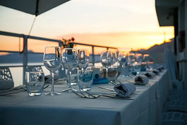 Long restaurant table set with wine glasses, folded napkins and cutlery on a waterfront terrace at sunset