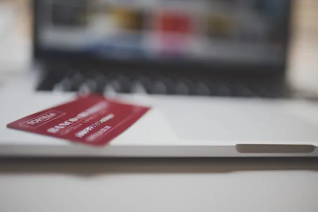 Red payment card resting on the edge of a silver laptop, with a blurred online store on the screen behind it