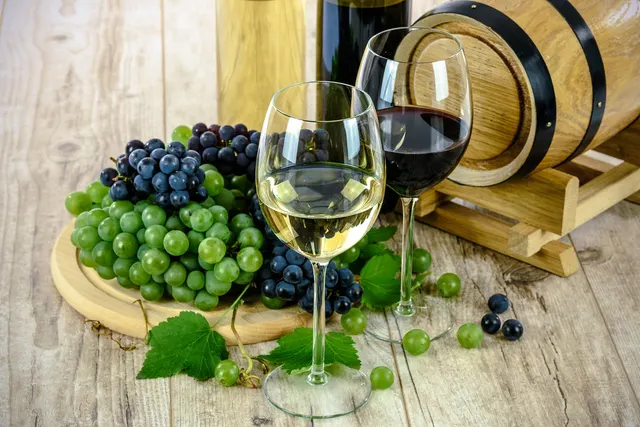 Glasses of white and red wine beside green and purple grape bunches, wine bottles and a small oak barrel on a wooden table