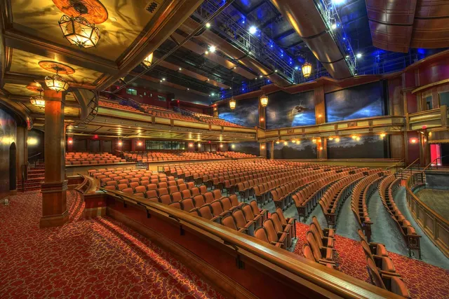 Empty theater auditorium with rows of orange seats, a curved balcony, warm hanging lanterns and blue stage lighting