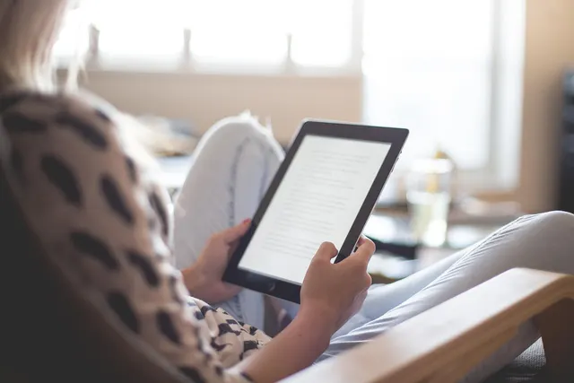 Woman in a leopard-print top relaxing in a wooden armchair, reading an article on a tablet by a bright window