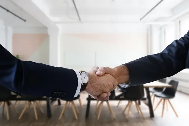 Two businessmen in dark suits shaking hands in front of an empty conference room with wooden chairs