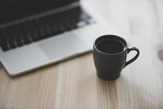 Silver laptop beside a dark gray mug of black coffee on a light wooden desk, keyboard softly out of focus