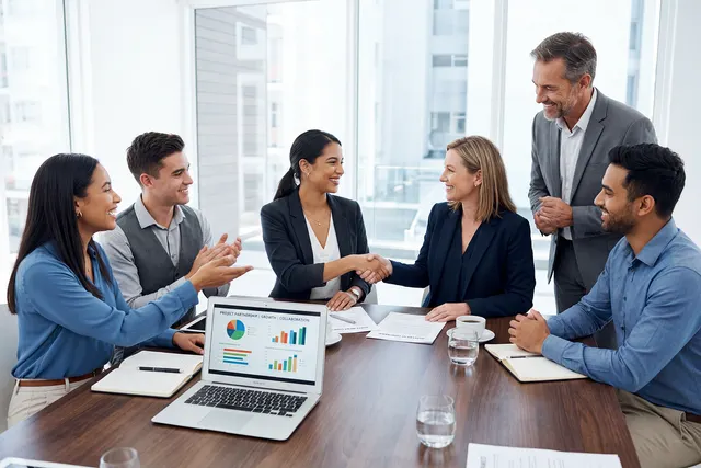 Smiling colleagues around a conference table applaud as two women shake hands beside a laptop showing charts