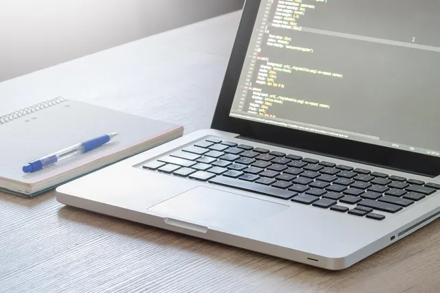 Laptop showing lines of code on screen beside a spiral notebook and blue pen on a wooden desk