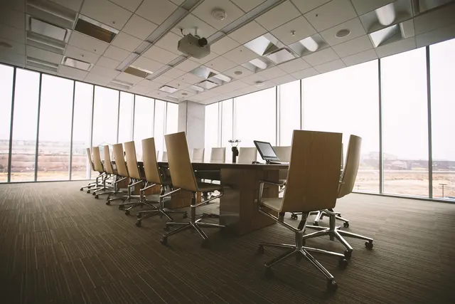 Empty conference room with a long wooden table, beige office chairs, a laptop and floor-to-ceiling windows