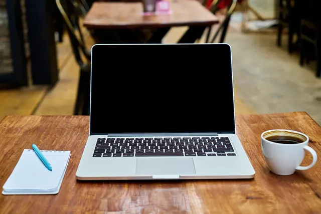 Open laptop with a blank screen on a wooden café table beside a notepad, blue pen and cup of black coffee