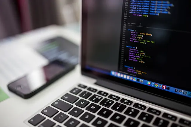 Close-up of a laptop showing CSS code in a dark editor, with a smartphone blurred on the desk beside it