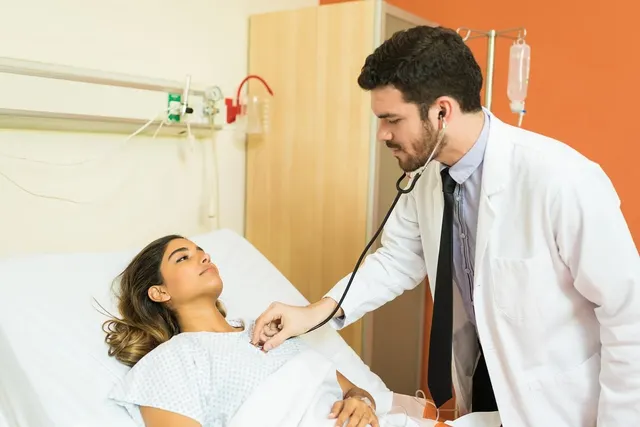 Doctor in a white coat listens with a stethoscope to a patient in a gown lying in a hospital bed beside an IV pole