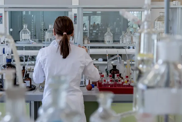 Scientist in a white lab coat, seen from behind, working at a lab bench lined with glass bottles and reagent flasks
