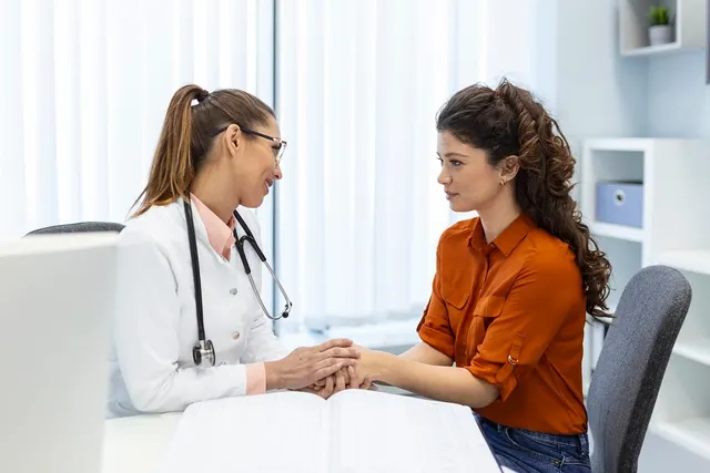 Doctor in white coat with stethoscope holds the hands of a woman in an orange blouse across a desk in a bright clinic office