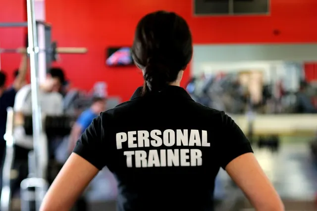 Personal trainer in a black shirt seen from behind, overlooking a busy gym floor with red walls and exercise equipment
