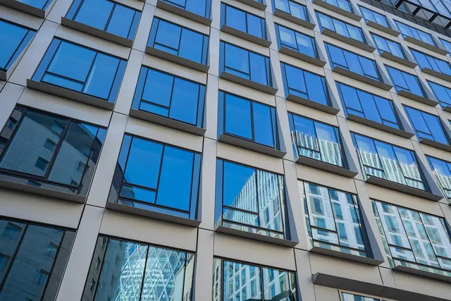 Low-angle view of a modern office building facade with rows of blue reflective windows mirroring nearby towers