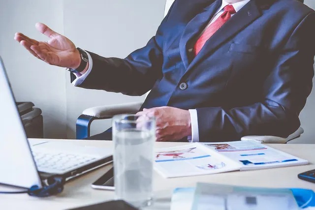 Businessman in navy suit and red tie gesturing at a desk with laptop, glass of water, phone and printed reports