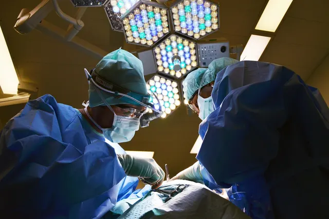 Surgeons in blue gowns, caps, masks and magnifying loupes operating beneath bright surgical lights in an operating room