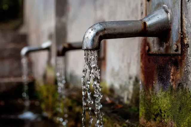 Water pouring from a row of weathered metal spouts on a mossy stone wall, with rust stains around the nearest pipe