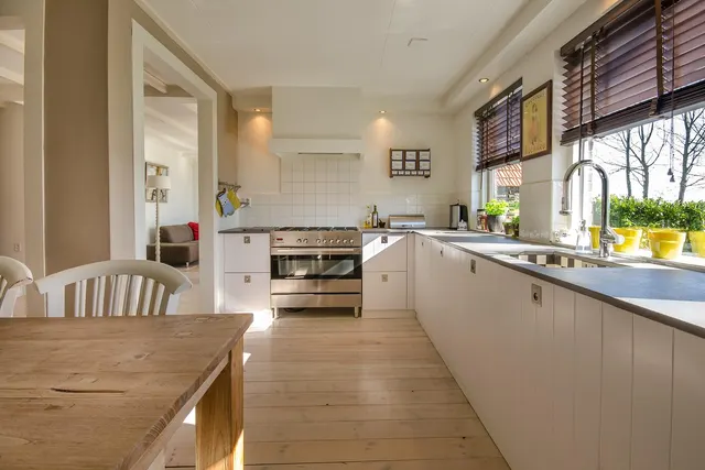 Sunlit home kitchen with white cabinets, a stainless steel stove, a window-side sink with chrome faucet, and a wooden table