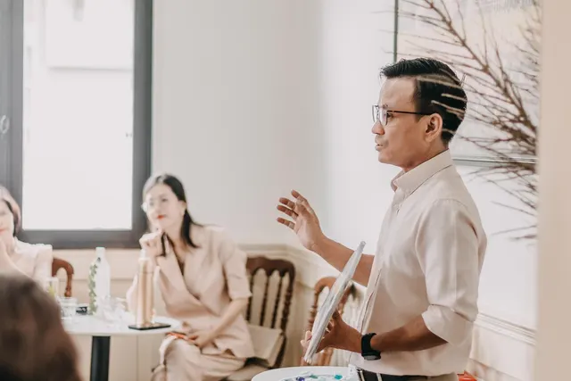 Man in glasses holding a tablet and gesturing while presenting to women seated at tables in a bright meeting room