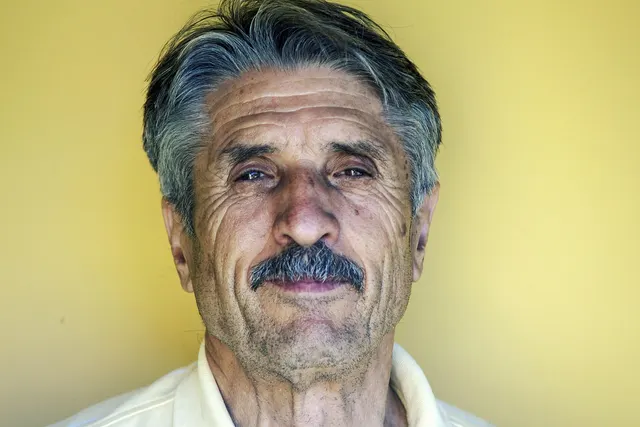 Close-up portrait of an older man with gray hair and a mustache in a white collared shirt against a yellow wall