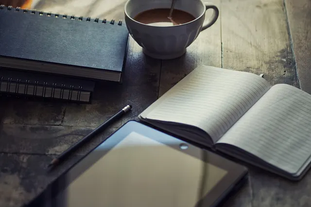 Tablet, open lined notebook, spiral notebooks, pencil and a cup of coffee on a rustic wooden table