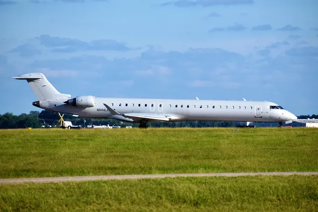 All-white regional jet with no airline livery taxiing past a grassy airfield under a blue, partly cloudy sky