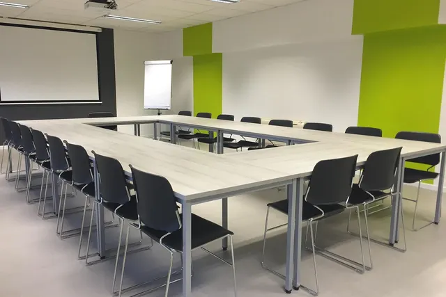 Empty conference room with black chairs around a U-shaped table, projector screen, flip chart and lime green wall accents