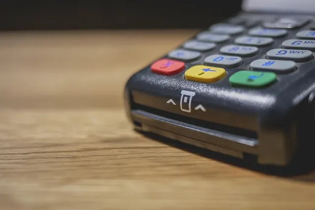 Close-up of a black card payment terminal with keypad and colored buttons, showing the card insert slot, on a wooden table