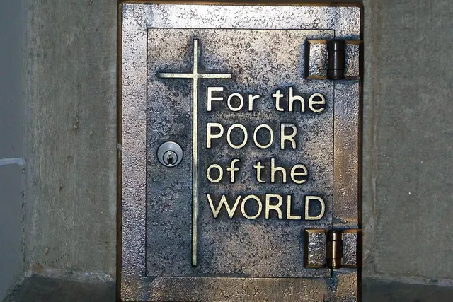 Bronze church donation box with a cross and the words "For the POOR of the WORLD" mounted on a stone wall