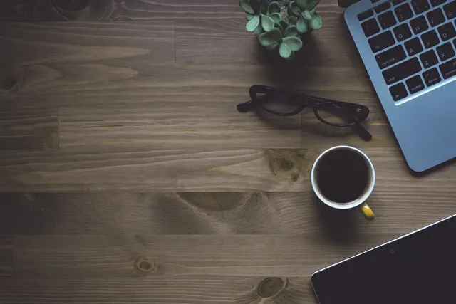 Overhead view of a wooden desk with a laptop, tablet, black glasses, a cup of black coffee and a small succulent
