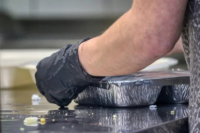 Gloved hand packing foil takeout containers on a stainless steel restaurant counter for a delivery order