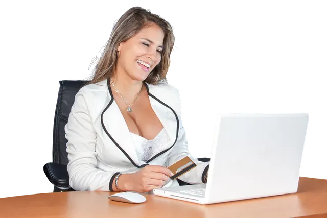Smiling woman in a white blazer at a wooden desk holding a payment card while using a white laptop, black background