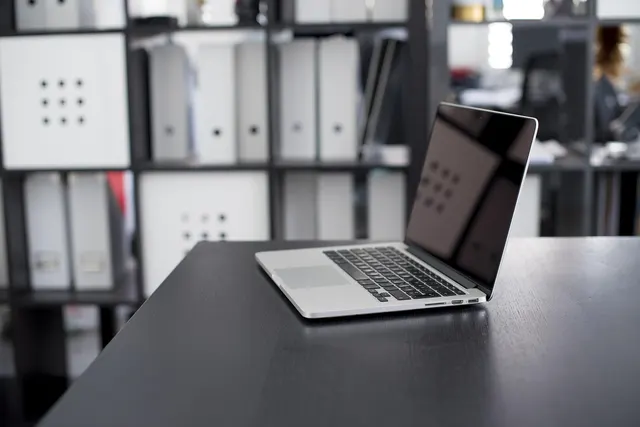 Open silver laptop on a dark desk in an office with shelves of white binders and storage boxes behind it