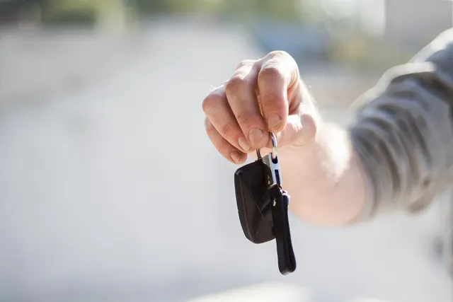 Hand in a gray sleeve holding out a black car key fob on a ring against a blurred, sunlit outdoor background