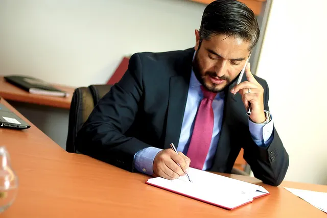 Bearded professional in a dark suit and pink tie talks on a phone while writing in a notebook at an office desk