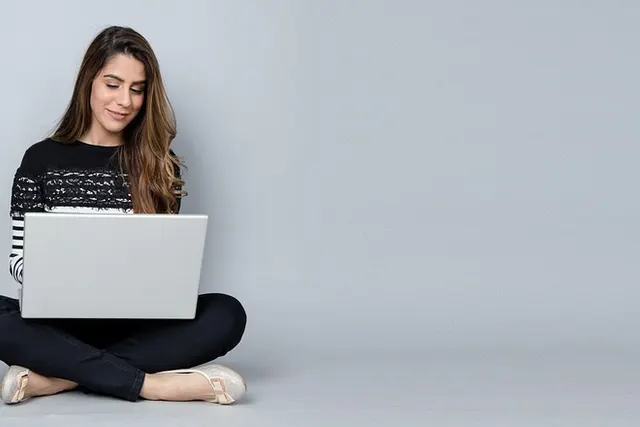 Smiling woman with long brown hair sits cross-legged on the floor using a silver laptop against a plain gray backdrop